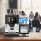 OBL3 Kalea Plus coffee machine with three bean hoppers and milk fridge on a wooden table in an office meeting room with people in the background