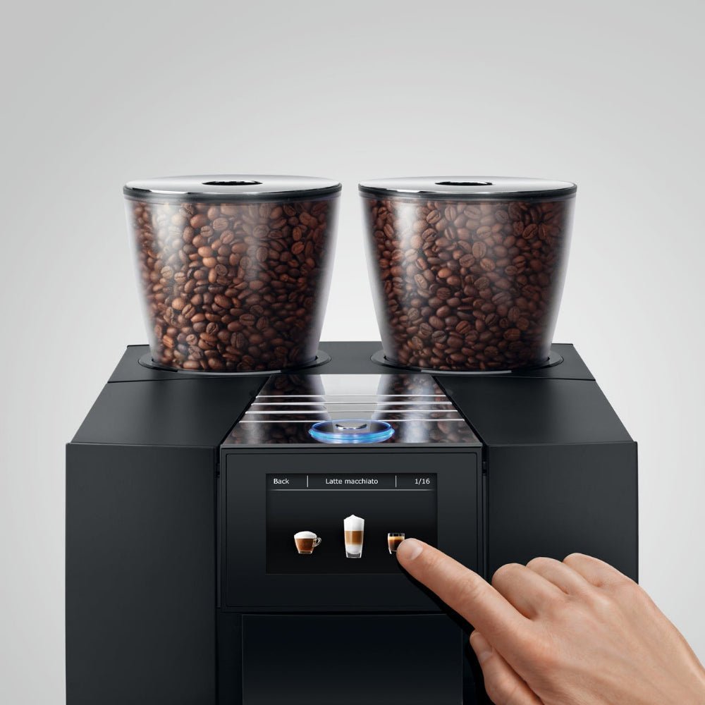 Top view of OBL2 Jura Giga X8C coffee machine with dual bean hoppers and a hand selecting a drink on the touch control panel on a light gray background