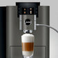 Close-up of OBM3  Jura X10 coffee machine spout dispensing a layered latte into a glass with milk foam on a dark grey background