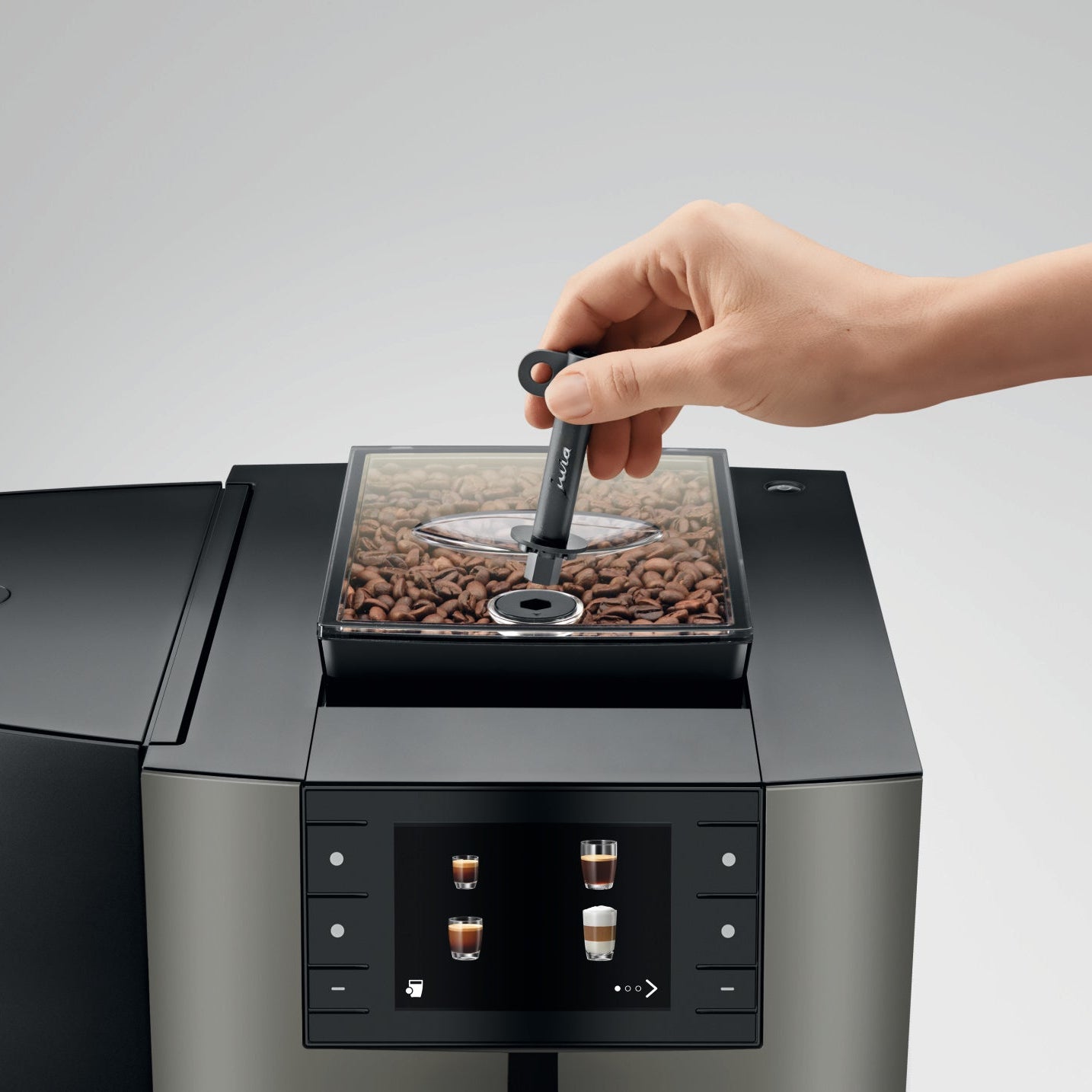 Hand placing a scoop into OBM3 Jura X10 coffee machine bean hopper with clear container filled with coffee beans on light gray background