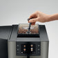 Hand placing a scoop into OBM3 Jura X10 coffee machine bean hopper with clear container filled with coffee beans on light gray background